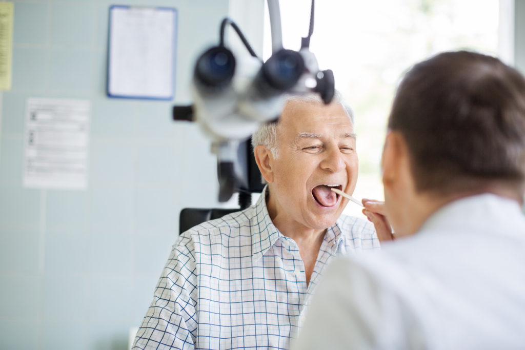 Senior man opening his mouth for the doctor to look in his throat. Male doctor examining sore throat of senior patient in clinic.